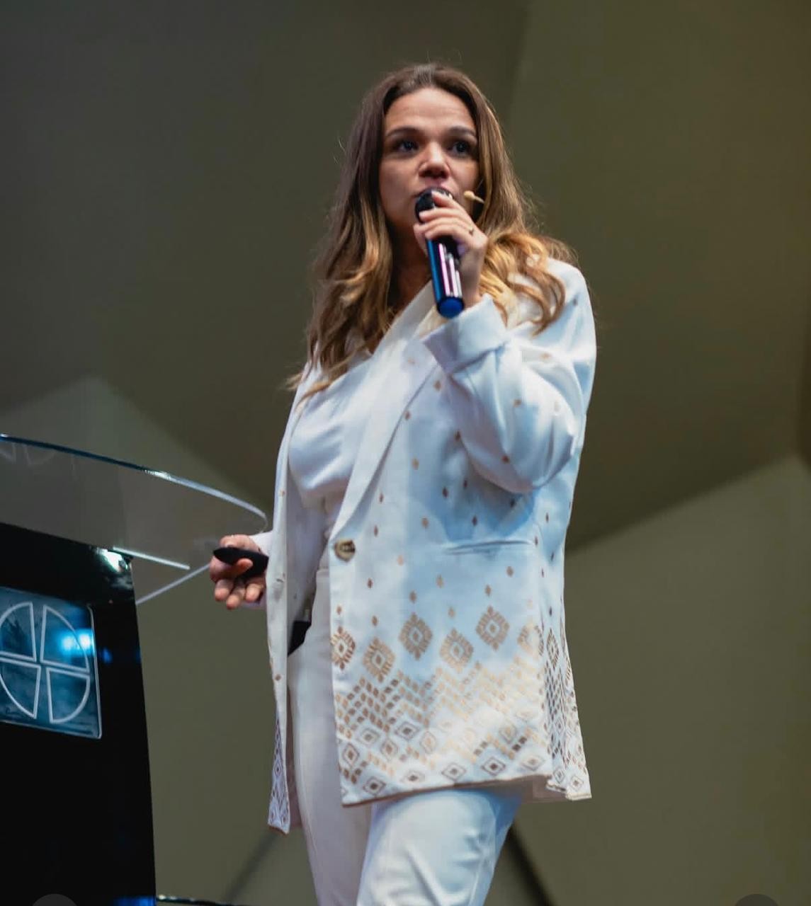 Dra. Aline Lago palestrando ao microfone, vestindo blazer branco bordado, durante apresentação sobre autenticidade